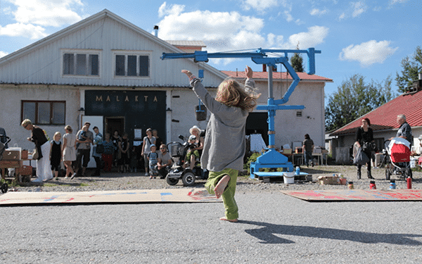 une enfant saute devant une grue bleu, située devant une facade avec écrit "malakta"